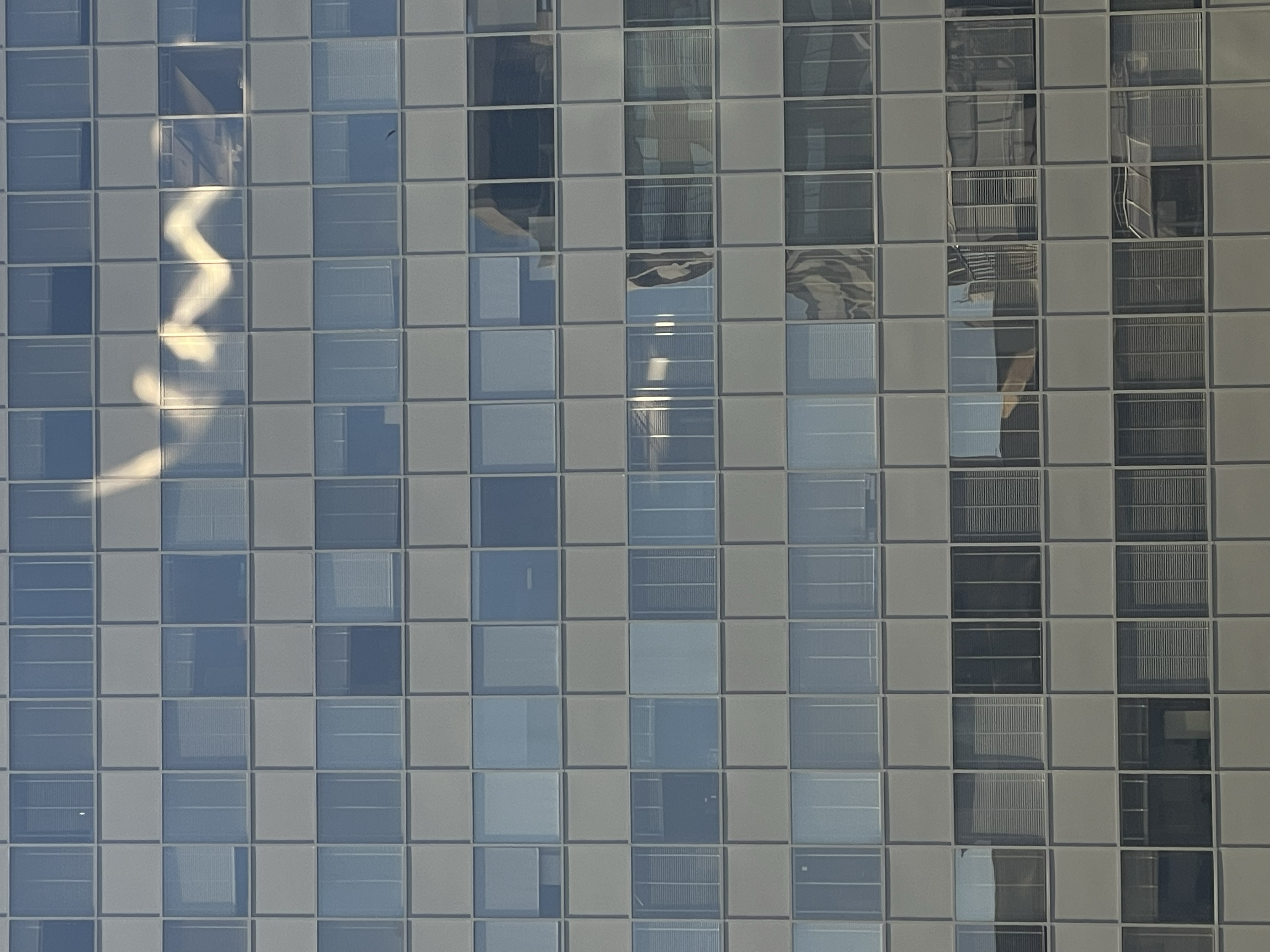 A glass office building fills the frame. Rows of square windows reflect the blue sky and surrounding buildings. A bright zigzag shape reflects near the top of the frame.